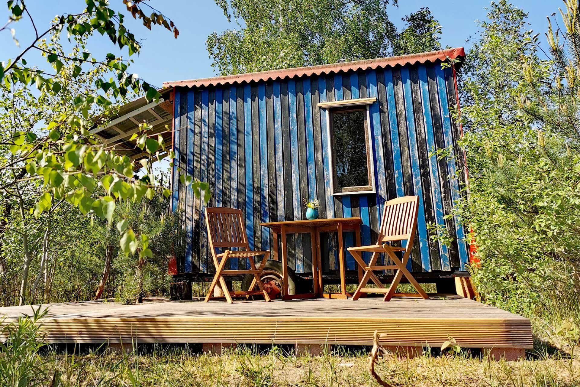Blau gestrichener Zirkuswagen mit Veranda und Stühlen am Waldrand